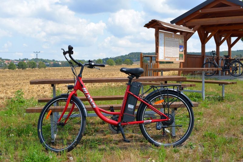 červený elektrobicykel HECHT RUNNIS opretý o lavičku vedľa cyklochodníka