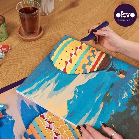 RUKA NANÁŠEJÍCÍ AKRYLOVOU BARVU NA NEBE V OČÍSLOVANÉM POLI PLÁTNA