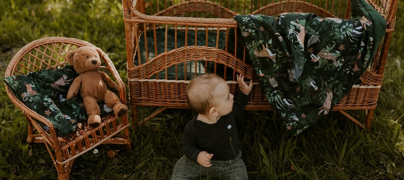 Miminko sedí u postýlky ze které vykukuje souprava postýlky ve vzoru Woodland značky Makaszka.